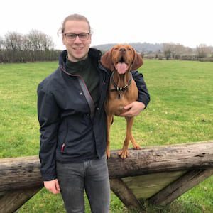 A man standing in a grassy field holding a brown dog while smiling at the camera.
